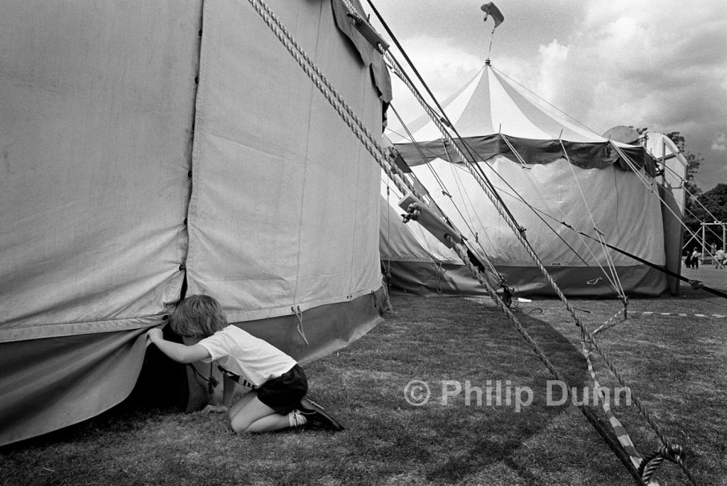 A small boy lifts the canvas of a circus tent to look inside