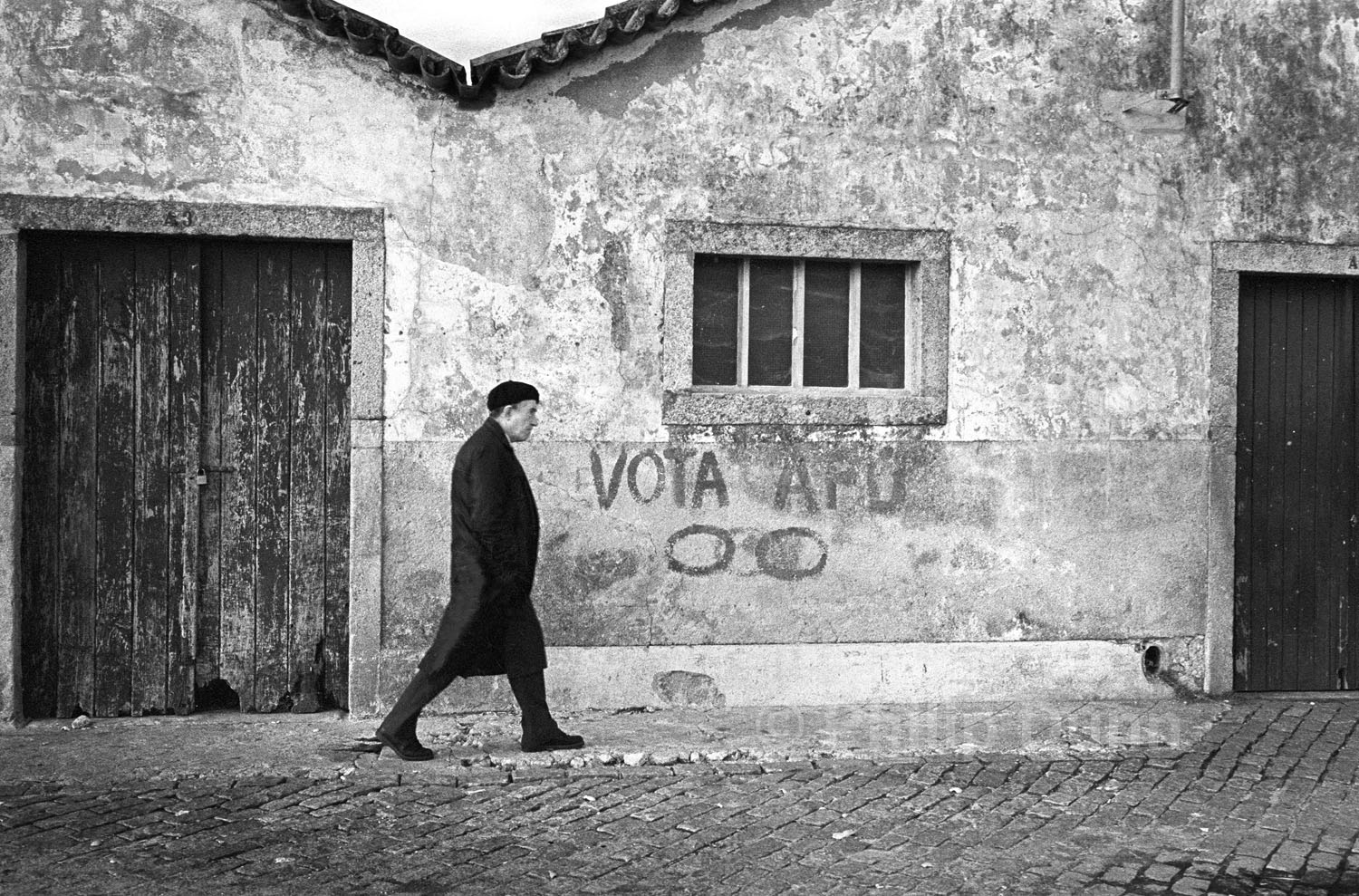 black and white image of man in black coat and beret walking past graffiti on Portuguese wal