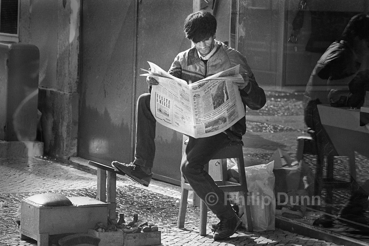 Shoe shine man reading paper, Portugal. black and white image