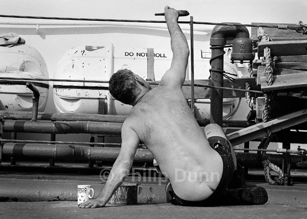Man paints ship's rail while displaying the cleavage of his buttocks