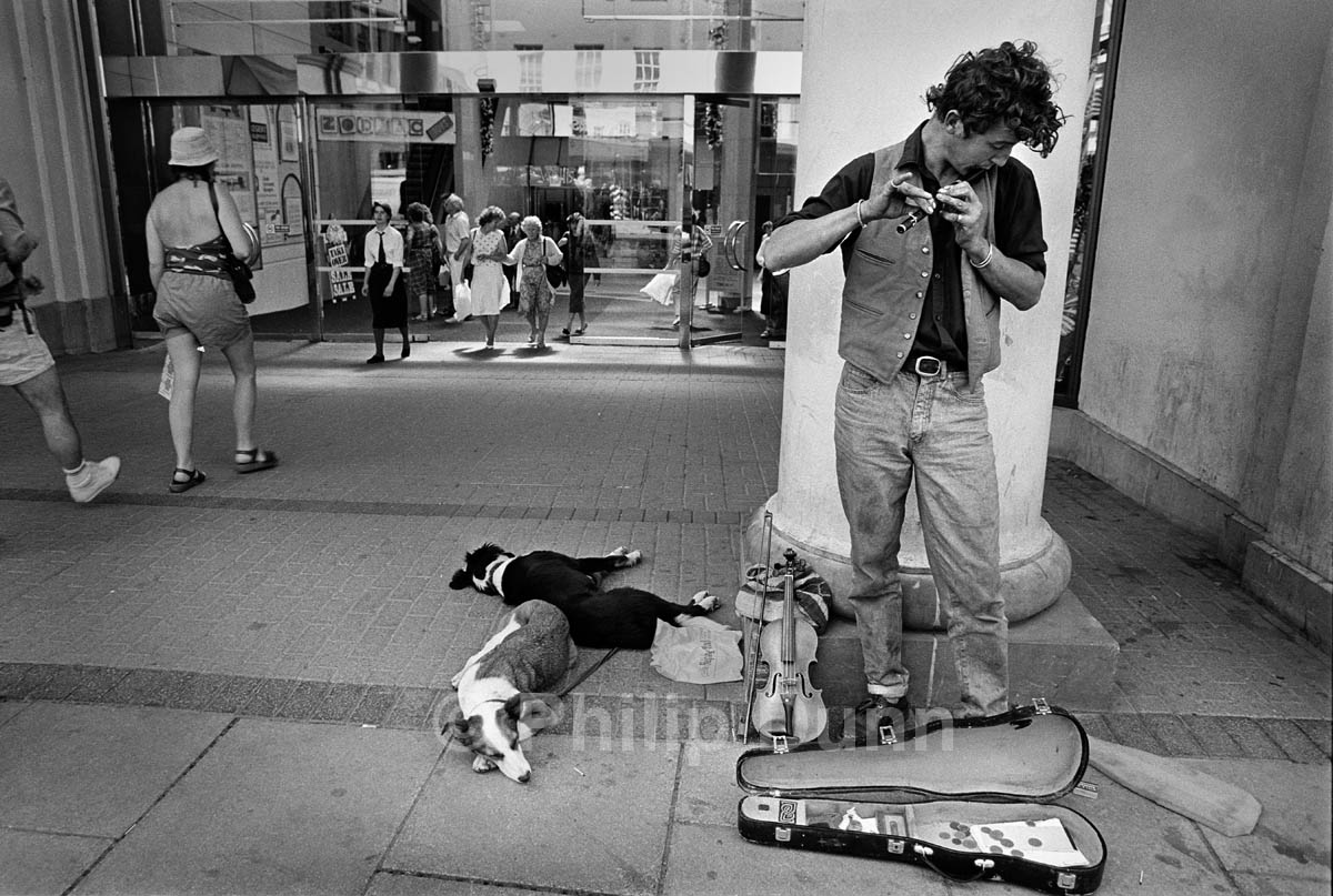 Street photography. A busker plays tin whistle outside a shopping centre while his dogs sleep