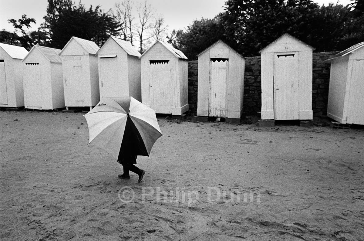 Child with large umbrella walks along beach, Bathing huts behind