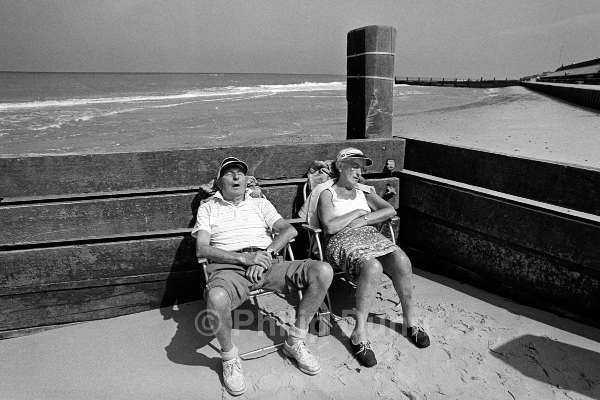 Elderly couple snooze on a Norfolk beach in shelter of a breakwater