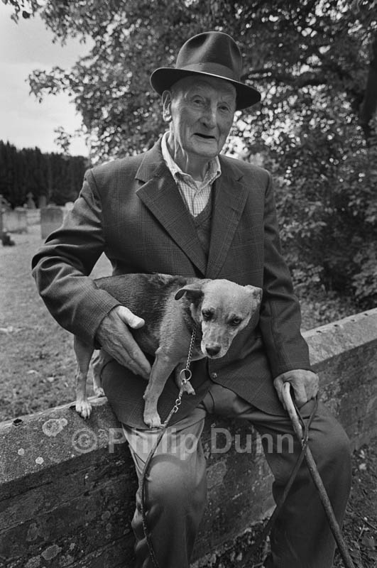 An old man sits on a churchyard wall with his dog.  Harrow Way, Hampshire