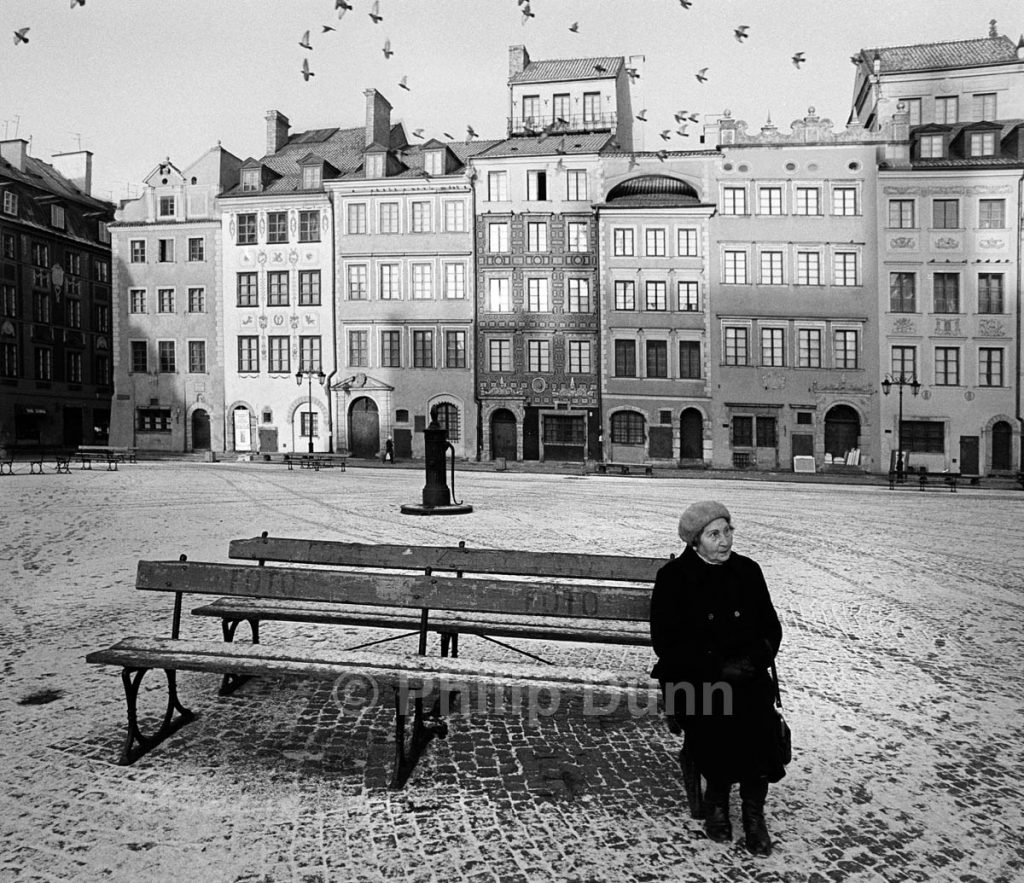 An old woman in hat sits on a bench in the snow Starigrad Square, Poland in Winter