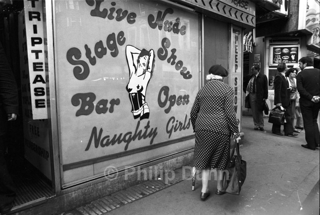 Old lady in beret passes strip club in Soho, London