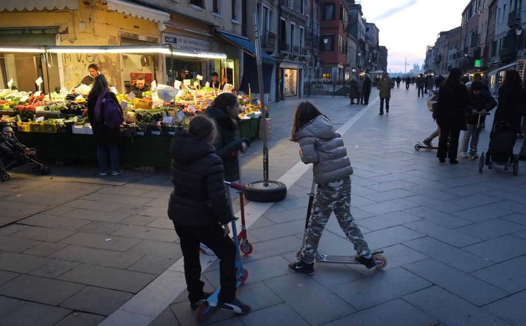 Street Photography myth and fact. Young girls on scooters in a Venice street at dusk. Behind the girls is a vegetable stall lit by light bulbs. The street goes away into the distance towards the Grand Canal