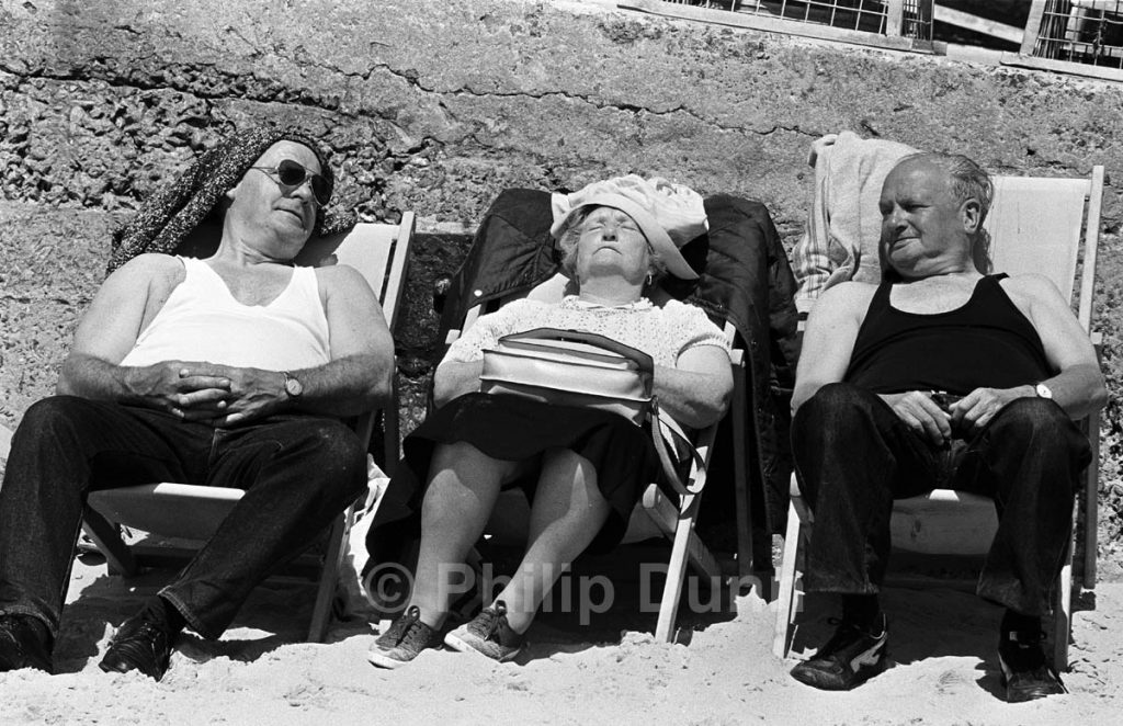 Two old men look at a sleeping old lady in deckchairs, Tenby Wales