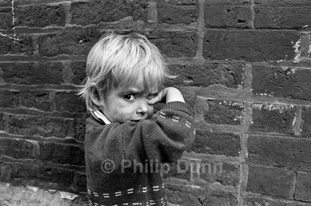 portrait of a small fair-haired boy with a dirty face, standing against a brick wall while rubbing his eye with his thumb